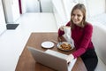 Young girl freelancer sits in a cafe with a laptop, drinks coffee and has breakfast, a student eats in the morning with a computer Royalty Free Stock Photo