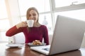 Young girl freelancer sits in a cafe with a laptop, drinks coffee and has breakfast, a student eats in the morning with a computer Royalty Free Stock Photo