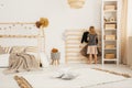 Young girl hanging clothes on wooden hanger in white Nordic style bedroom interior with home-shape bed, two pillows placed on car