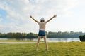 Young girl with her back in hat on the river bank, spread her hands in the sides, looks at the setting sun. Enjoys and meditates