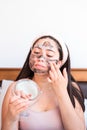 Young girl on her bed, applying facial clay mask. Skin care at home.