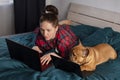 Young girl and her dog french bulldog working in bed at a laptop during quarantine Royalty Free Stock Photo