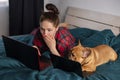 Young girl and her dog french bulldog working in bed at a laptop during quarantine Royalty Free Stock Photo