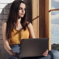 Young girl holding laptop while sitting on window sill and looking out with sad face impression Royalty Free Stock Photo