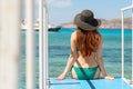 A young girl with a hourglass figure sits on a pier and looks into the sea. wearing a green swimsuit and a black hat. rear view.