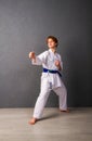 A young girl karateka in white kimono and a blue belt trains and performs a set of exercises against a gray wall Royalty Free Stock Photo