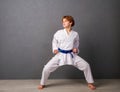 A young girl karateka in white kimono and a blue belt trains and performs a set of exercises against a gray wall Royalty Free Stock Photo