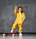 Young girl kid in yellow hooded jumpsuit with hood over her head and white sneakers stands on red skateboard and show peace sign