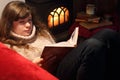 Young girl lady, woman sitting on sofa near wood stove with fire and her long brown hair lie on sweater and reading book Royalty Free Stock Photo