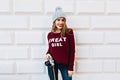 Young girl with long hair in marsala sweater on grey wall background. She wears knitted hat, holds camera and smiling.