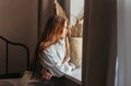 A young girl with long hair sitting in bed after waking up looking in window at city, indoors at home early morning, lifestyle Royalty Free Stock Photo