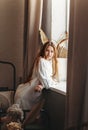 A young girl with long hair sitting in bed after waking up looking in window at city, indoors at home early morning, lifestyle Royalty Free Stock Photo