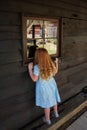 A young girl with long red hair and a blue dress and upstreched arms looks curiously through a window of an old wooden building Royalty Free Stock Photo