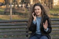 Young girl with long wavy brown hair sits on bench in park talks on phone and gesticulating. Woman in autumn park Royalty Free Stock Photo