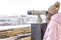 A young girl looks through coin-operated binoculars on the observation deck overlooking the city from a height at sunset. Winter,