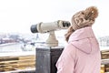 A young girl looks through coin-operated binoculars on the observation deck overlooking the city from a height at sunset. Winter,