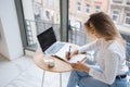 Young girl is making notes in cafeteria. Cup of coffee, water glass and laptop on freelancer`s desk. Royalty Free Stock Photo