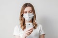 Young girl in a medical mask calls on the phone and looking at a cell phone isolated over light grey background, ordering goods,