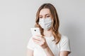 Young girl in a medical mask reads a news in her mobile phone isolated over grey background. Woman orders goods, ambulance call,