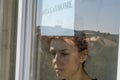 A young girl in a medical mask stands at the window with a sign saying stayathome