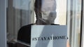 A young girl in a medical mask stands at the window with a sign saying stayathome