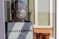 A young girl in a medical mask stands at the window with a sign saying stayathome