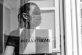 A young girl in a medical mask stands at the window with a sign saying stayathome