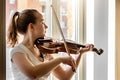 A young girl, a musician, plays the violin on the background of a window