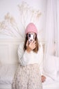 A young girl with a phone in her hands covers her face in a bright room. Fashionable knitted hats autumn-winter.