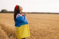 Young girl prays in the wheat field wearing Ukrainian national embroidered shirt with flower red wreath and holds flag at sunset y Royalty Free Stock Photo