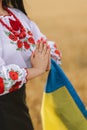 Young girl prays in the wheat field wearing Ukrainian national embroidered shirt with flower red wreath and holds flag at sunset y Royalty Free Stock Photo