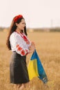 Young girl prays in the wheat field wearing Ukrainian national embroidered shirt with flower red wreath and holds flag at sunset y Royalty Free Stock Photo