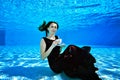 A young girl in a purple dress is sitting underwater at the bottom of the pool, holding a white Cup in her hand, looking at me Royalty Free Stock Photo