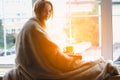 Young girl reading book and drinking coffee sitting next to the window wrapped in warm comfy blanket, Royalty Free Stock Photo