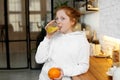 Young girl with red curly hair drinks orange juice standing in the kitchen, healthy food for baby Royalty Free Stock Photo