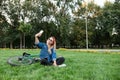 Young girl sits in the grass in park and takes selfie on a smartphone. Happy beautiful girl in stylish clothes and sunglasses sits Royalty Free Stock Photo