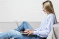 A young girl sits on a gray sofa against a white brick wall with a laptop on her lap. Profile view. Distance learning, distant