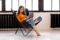 A young girl sits at home in a stylish black leather chair, holds a cup of coffee and a tablet in her hand Royalty Free Stock Photo