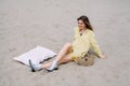 A young girl sits on the sand, on the beach, rests and smiles, next to it lies a pillow and a basket with a bottle of champagne