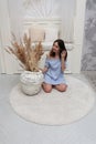 A young girl is sitting on the floor on a carpet with a vase. Pretty young woman in a blue dress looks into the frame in