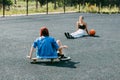 A young girl is sitting on a skateboard outdoors on a basketball court with her basketball player friend