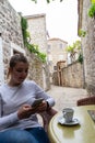 Young girl smiling typing a message in smart phone while take a coffee in a terrace of a stone narrow street in Budva, Montenegro Royalty Free Stock Photo