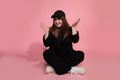 Young girl in sportswear and baseball cap sits on floor on pink background in studio. She spread her arms, emotions, joys,