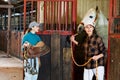 Young girl stable worker ready to saddle horse while Asian woman leading out of stall