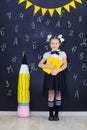 Young girl standing in front of chalkboard brick wall with letters on it wearing school uniform with huge pensil yellow color