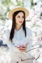 Young lovely girl in summer hat standing near Magnolia blossoming flowers, stands in near magnolia tree Royalty Free Stock Photo