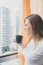 A young girl is standing by the window with a mug of coffee in her hands and looks out the window at the city landscap Royalty Free Stock Photo