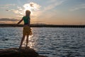 Young girl stood on rock at edge of water looking at sunset