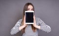 A young girl in a striped jacket covers her face with a tablet Royalty Free Stock Photo