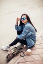 Young girl student is sitting on the pavement. Wear jeans and drink coffee. Young beautiful model girl in sunglasses smiles Royalty Free Stock Photo